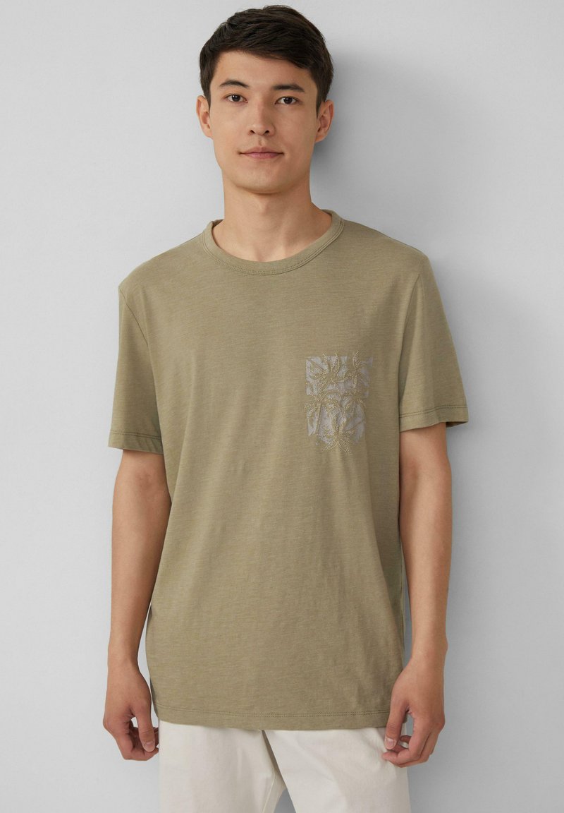 Oliwkowa bawełniana koszulka z krótkim rękawem i okrągłym dekoltem, z wzorzystą kieszenią po lewej stronie. Gładka tekstura.