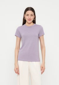 Νεαρή γυναίκα με ίσια μακριά μαλλιά φοράει χρυσούς κρίκους στα αυτιά, ένα κοντομάνικο μπλουζάκι λεβαντίνης και λευκό παντελόνι με φαρδιά μπατζάκια, στέκεται, κοιτώντας προς τα εμπρός.