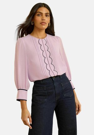 Hellviolette Bluse mit gezacktem Vorderteil, schwarzer Paspelierung entlang der Ärmel und elastischen, aufgepufften Ärmeln; kombiniert mit hoch taillierten dunklen Jeans.