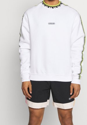 Persoon draagt een witte Adidas-trui met zwarte en neongele stikdetails en zwarte Adidas-short met beige boorden.