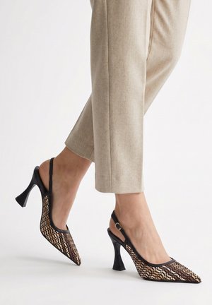 Piedi con pantaloni beige e décolleté slingback con motivo, tacco nero e punta appuntita su sfondo bianco.