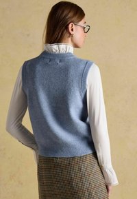 Naine kannab helesinist kootud sviitrit valge triikitud kraega pluusi peal, mustriga houndstooth seeliku all, seistes seljaga beige tausta poole.