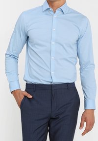 Camicia a maniche lunghe di colore blu chiaro con colletto a punte, frontale abbottonato e tessuto liscio, indossata con pantaloni sartoriali blu navy.