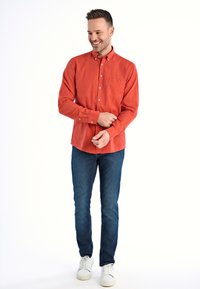 Czerwona koszula na guziki z kieszenią z przodu, w połączeniu z ciemnoniebieskimi dżinsami i białymi trampkami. Posiada długie rękawy i luźny krój.