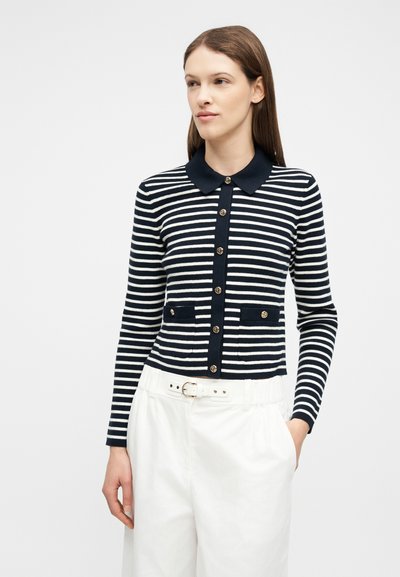 Femme portant un pull boutonné à rayures bleu marine et blanc avec col, et un pantalon blanc avec ceinture, debout avec une main dans la poche.