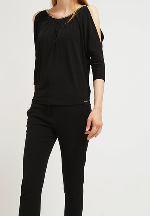 Femme portant un haut noir à épaules dénudées avec un détail de chaîne dorée et un pantalon noir ajusté, debout devant un fond uni.