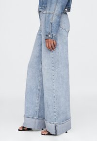 Svetlomodré široké rifle s vysokým pásom, veľkými manžetami a bočnými vreckami. Tkanina má jemnú textúru s viditeľným prešívaním.