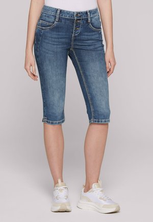 Slim Fit blaue Denim-Capri-Jeans mit Knopfverschluss, die unterhalb der Taille sitzen, kombiniert mit weißen Sneakers und einem weißen Top.