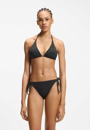 Femme aux cheveux tressés portant un bikini noir texturé avec un haut à col halter et un bas noué sur les côtés, debout sur un fond blanc.