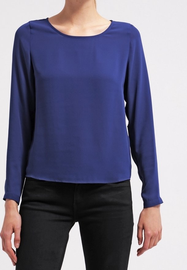 Personne portant un chemisier bleu roi à manches longues et un pantalon noir, debout devant un fond clair uni.