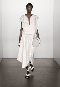 White sleeveless top with a deep V-neck, paired with a flowing white skirt. Model carries a woven white bag and wears white strappy sandals.