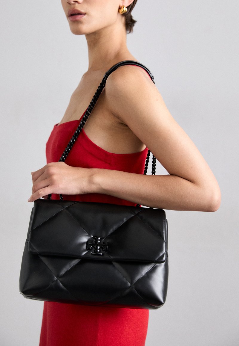 Black quilted leather handbag with a fold-over flap and logo clasp, featuring a dual textured strap and smooth finish, held beside a model in a red dress.