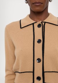 Beige cardigan med krage, svarte kantdetaljer og fem svarte knapper foran. Myk tekstur, tettsittende design.