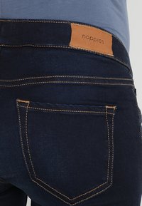 Vaqueros azul oscuro con una textura suave, con un parche de cuero marrón claro que lleva la marca "noppies", costuras amarillas y bolsillos traseros.