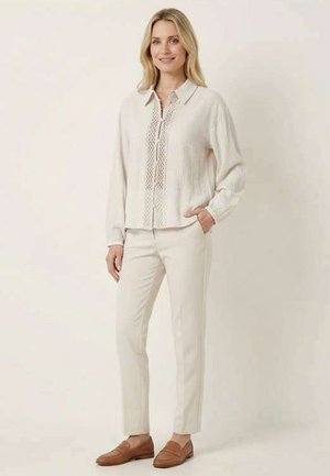 Femme debout en blouse crème avec détail en dentelle, pantalon assorti et mocassins marron devant un fond uni.