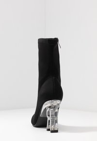 Bottine noire avec tige en tissu ajusté et talon bloc transparent, exposée sur une surface blanche contre un fond clair uni.