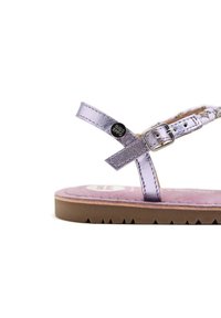 Sándal metálico en color lila con una correa de tobillo con hebilla, plantilla de textura suave y suela de goma marrón con un patrón en zigzag.