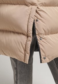 Beige puffjakke med en quiltet design, metalliske trykknapper og lynlåsåbning. Blød, polstret tekstur og en kontrasterende sort foring.