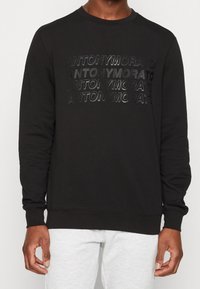 Sudadera negra con un logo texturizado impreso "ANTONYMORATO" en diferentes opacidades. Cuello redondo, mangas largas y puños de canalé.