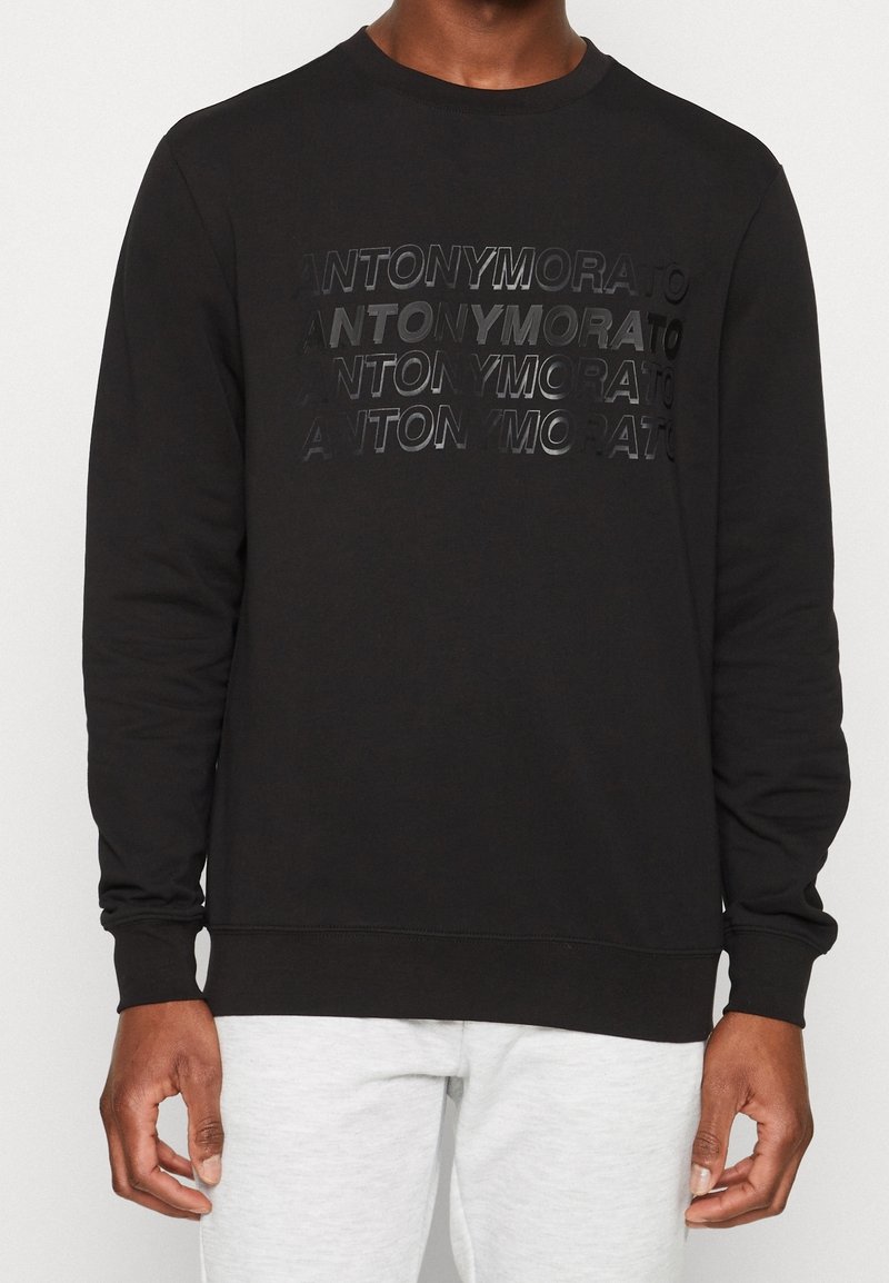Sudadera negra con un logo texturizado impreso "ANTONYMORATO" en diferentes opacidades. Cuello redondo, mangas largas y puños de canalé.
