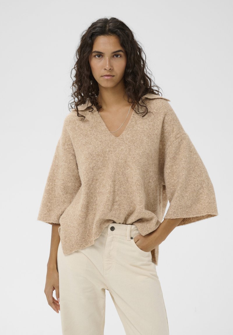 Femme aux cheveux bouclés portant un pull ample col V beige et un pantalon crème, debout avec une main dans la poche sur un fond uni.