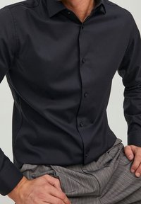 Camisa negra de botones, hecha de una tela suave, con un cuello estructurado, mangas largas y un ligero brillo; combinada con pantalones a rayas.