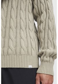 Pull en tricot torsadé beige clair avec des poignets et un ourlet côtelés, présentant des motifs torsadés bien visibles et une petite étiquette avec logo en bas.