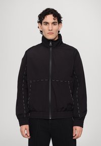 Schwarze Windjacke mit hohem Kragen, Reißverschluss und weißen Nähten. Leichtes Material und lockere Passform für Komfort.