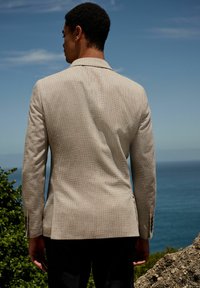 Beige textured blazer viewed from the back, featuring a tailored fit, notch lapels, and buttoned cuffs, against a coastal background.