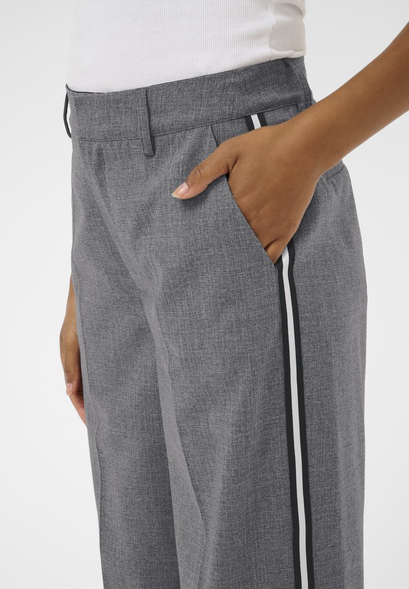 Un pantalon gris avec un tissu texturé, coupe ample, poches latérales et rayures verticales noires et blanches sur les côtés.