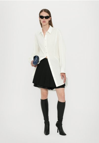 Chemise blanche surdimensionnée à boutons, jupe mini plissée noire, bottes à talons noires montantes, et petit sac à main en textile bleu.