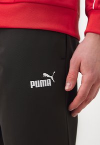 Čierne športové nohavice s hladkou textúrou, so bielym logom Puma na stehne. Nosič má ruku opretú o nohavice.