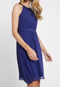 Bezramenn dress s délkou po kolena v hlubokém modrém šifonu, s perlovým výstřihem a nařaseným pasem. Diskrétní detaily ve tvaru klíčové dírky na hrudi.