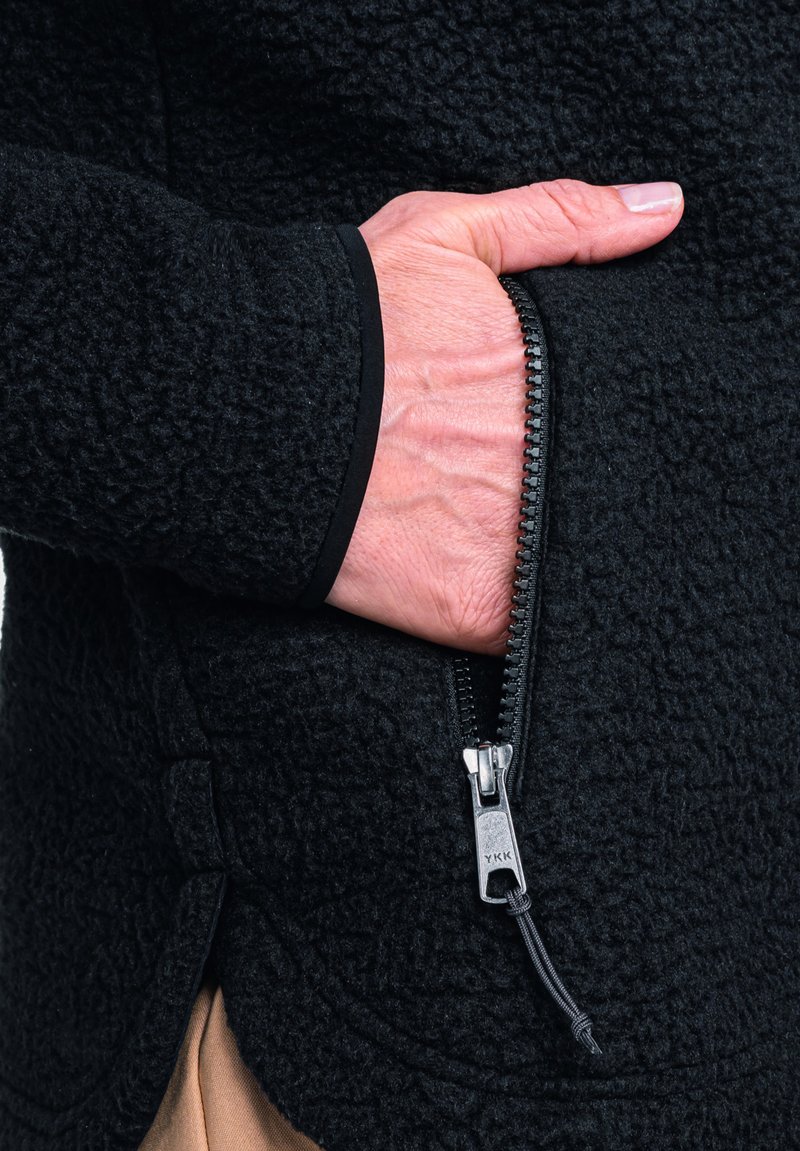 Giacca in pile nera con finitura testurizzata; presenta una tasca con cerniera e accessori YKK; primo piano di una mano che tiene la tasca.