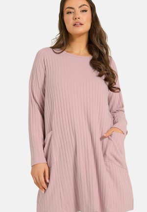 Frau trägt ein langärmeliges, geripptes, hellrosa Kleid mit Taschen, steht vor einem schlichten Hintergrund mit einer Hand in der Tasche.