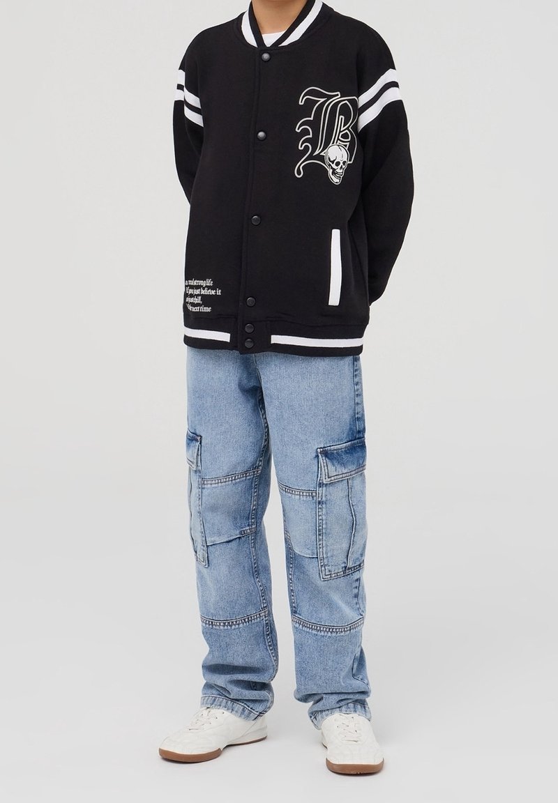 Personne portant une veste varsity noire avec un motif de crâne et la lettre B, un jean cargo bleu clair et des baskets blanches, debout devant un fond uni.