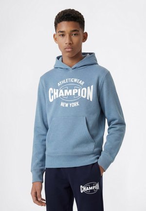 Nastolatek ubrany w niebieską bluzę Champion z kapturem oraz granatowe spodnie z logo Champion, stoi z jedną ręką w kieszeni na białym tle.