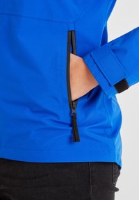 Hand in zipper pocket of blue jacket with adjustable Velcro cuff, worn with black pants.