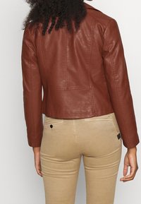 Kobieta w brązowej skórzanej kurtce i beżowych dopasowanych spodniach, pokazana od tyłu na jednolitym jasnym tle.