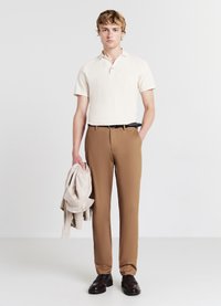 Cremfarbener Poloshirt mit kurzen Ärmeln, braune maßgeschneiderte Hose, schwarzer Gürtel, hält eine hellgraue Jacke und steht vor einem schlichten Hintergrund.