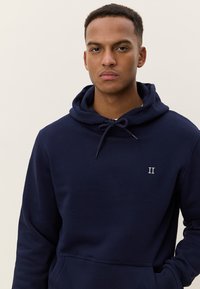 Tmavomodrá mikina s kapucňou a šnúrkami, predným vreckom a vyšitým označením "II" na hrudi. Jemný materiál a bežný strih. Hladká textúra.