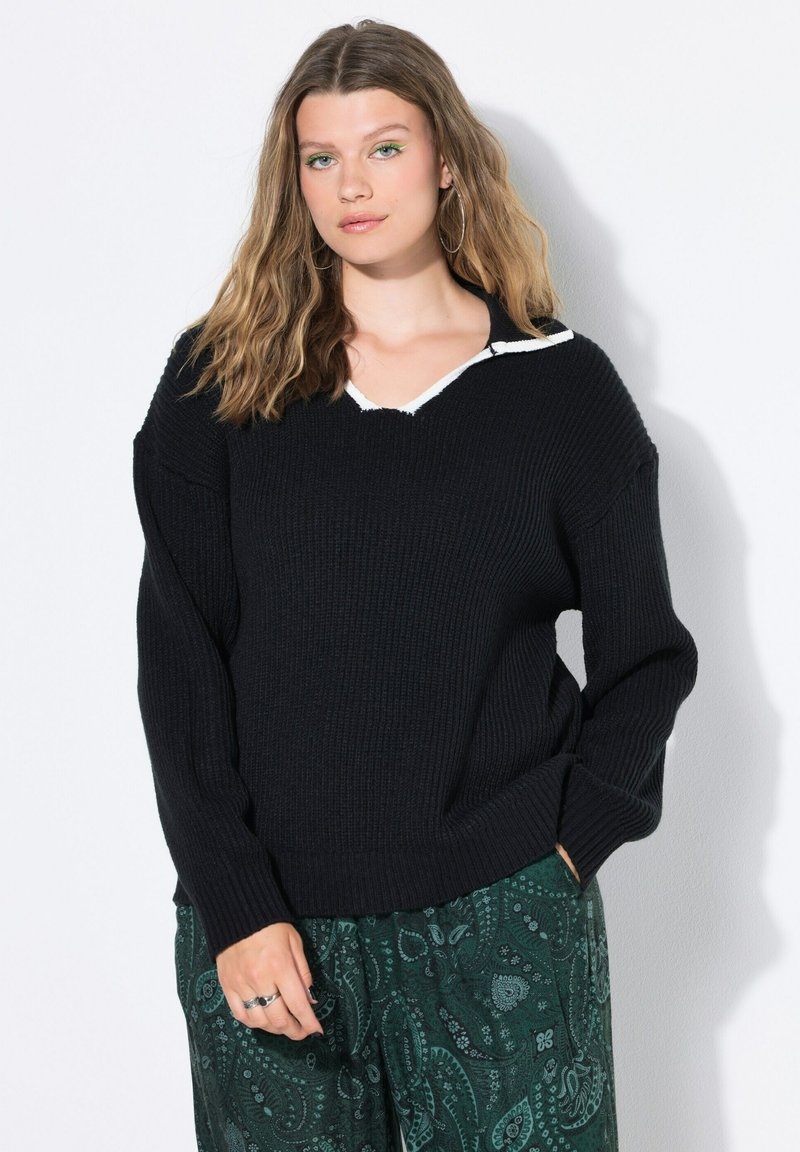 Vrouw met golvend blond haar, die oorringen draagt, een zwarte gebreide trui met witte kraagdetails, en groene paisley-patroon broeken op een witte achtergrond.