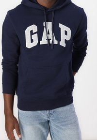 Sudadera azul marino de algodón con un bolsillo frontal. Presenta letras blancas "GAP" y cordones en la capucha. Ajuste casual.