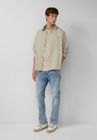 Beige overhemd met knopen, lichtgrijze t-shirt eronder, lichtblauwe spijkerbroek en witte sneakers met tan accenten. Eenvoudig ontwerp, casual pasvorm.