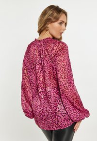 Blusa trasparente rosa con un motivo a stampa leopardata e dettagli dorati. Presenta maniche a sbuffo e un colletto arricciato. Tessuto morbido e leggero.
