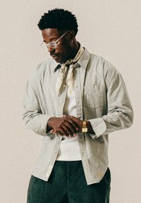 Mann mit lockigem Haar, der eine Brille trägt, gestreifte Hemd, weißes T-Shirt, Schal, goldene Uhr und Anhänger, nach unten schaut mit gefalteten Händen.