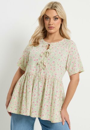 Blondynka w krótkim rękawku, bluzka w kwiatowy wzór z trzema wiązaniami z przodu oraz niebieskie dżinsy, stojąca na jednolitym tle.