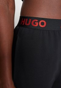 Črne kratke hlače z mehko teksturo in elastičnim pasom, na katerem je ime blagovne znamke "HUGO" napisano z rdečimi črkami ob vrhu.