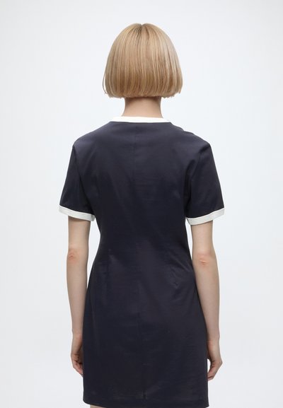 Personne avec une coiffure bob courte blonde portant une robe noire à manches courtes avec un liseré blanc, debout de dos devant un fond uni.