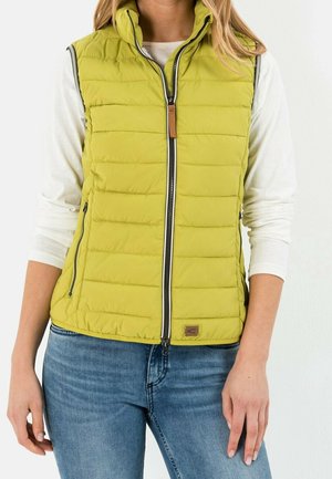 Femme portant un gilet matelassé sans manches vert citron sur un chemisier blanc à manches longues avec un jean bleu, debout devant un fond blanc.